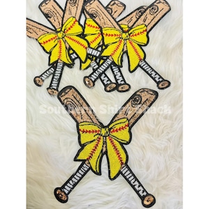 Pode incluir: Enfeites decorativos com tema de softball. A imagem apresenta vários bastões de beisebol cruzados com um laço amarelo e costuras vermelhas. Os bastões têm um design de grão de madeira marrom e os laços são adornados com lantejoulas amarelas. O fundo é branco.