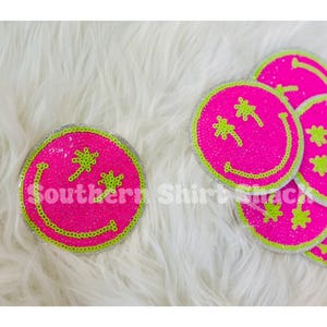 May include: Bright pink smiley face patches with green and silver accents. The patches feature a smiling face design with star-shaped eyes and the text "Southern Shirt Shack". These patches are likely for clothing or accessories.