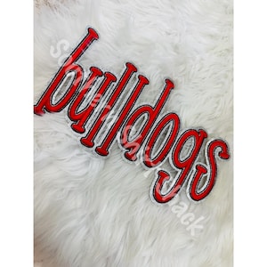Op de afbeelding: Een decoratieve patch met het woord Bulldogs in een gestileerd rood lettertype met een dikke zilveren glitterrand. De letters zijn verbonden in een cursieve schrijfstijl, waardoor een enkel samenhangend stuk ontstaat met een gedurfde uitstraling.