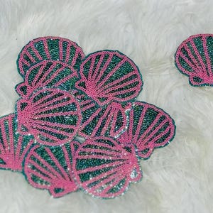 May include: A collection of seashell-shaped patches featuring a vibrant combination of bright pink and shimmering emerald green sequins. The patches are arranged in a cluster, with one individual seashell patch positioned separately to the right.