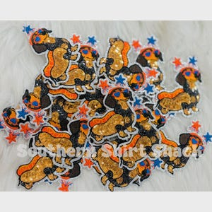 May include: A collection of sequined dachshund patches featuring dogs wearing heart-shaped sunglasses and party hats, accented with orange and blue star shapes. The patches are layered together, and the text Southern Shirt Shack is visible.