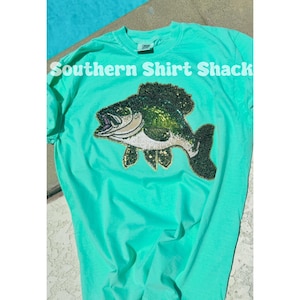 May include: A turquoise t-shirt with the text "Southern Shirt Shack" and a detailed fish design. The fish is embellished with sequins in shades of green, black, and silver, creating a shimmering effect. The shirt is made of a soft material.