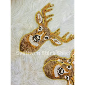 May include: Two sequined deer head decorations. The deer heads are brown, gold, and white, with detailed facial features and antlers. The text "Southern Shirt Shack" is visible at the bottom of the image.