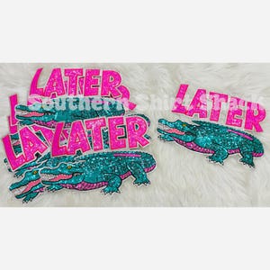 Può includere: Molteplici toppe termoadesive con una grafica di alligatore glitterato color verde acqua e verde, abbinata alla parola LATER in testo glitterato rosa brillante. I disegni sono sovrapposti e mostrano l'alligatore con la bocca aperta.
