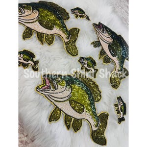 May include: A collection of six fish-shaped patches featuring green and cream sequins. The patches vary in size and depict largemouth bass with open mouths, detailed fins, and scales rendered in shimmering sequins. The text Southern Shirt Shack is visible.