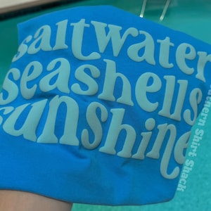 May include: A blue t-shirt with the words "saltwater seashells sunshine" in a light blue, puffy font. The words are stacked vertically. The text "Southern Shirt Shack" is printed vertically on the right side of the shirt.