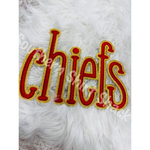 May include: A red and gold embroidered patch featuring the word chiefs in a stylized, rounded font. The letters have a bold red center with a thick, shimmering gold border, creating a distinct outline for each character in the word.