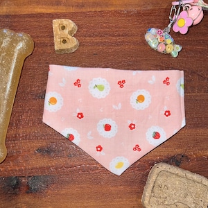 May include: A pink dog bandana with a repeating pattern of fruits and flowers. The bandana features strawberries, oranges, pears, and small red flowers on a light pink background. The bandana is triangular in shape and made of fabric.
