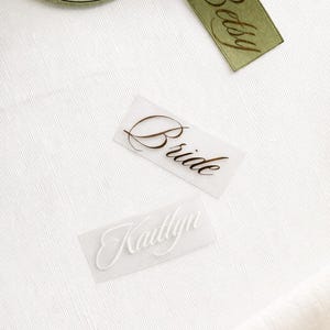 May include: A close-up shot features a green ribbon with the name "Betsy" printed in brown script. Two transparent labels display the words "Bride" and "Kaitlyn" in elegant fonts. The items rest on a white fabric surface.