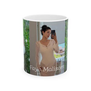 Puede incluir: Taza de cerámica blanca con una fotografía de una persona con un vestido blanco, sosteniendo un teléfono. La taza tiene el texto "Faye Malisom" impreso. El fondo incluye vegetación y una ventana.