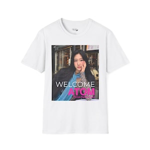 May include: White t-shirt featuring a photograph of a person with long black hair. The image on the shirt includes the words "WELCOME ATOM" in white, blue, and pink. The shirt has a crew neck and short sleeves.