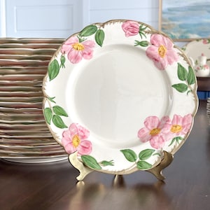 May include: A white ceramic dinner plate featuring a scalloped edge and a hand-painted floral pattern of pink wild roses with yellow centers and green leaves. The plate is displayed on a gold-toned metal stand in front of a stack of matching plates.