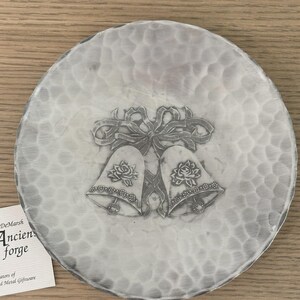 May include: A round, hammered metal plate with a decorative design. The design features two bells with roses and a ribbon. The plate has a textured surface and a silver color. The plate is a wedding or anniversary gift.