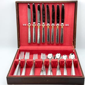 May include: A flatware set in a wooden storage box with a red velvet interior. The set includes eight stainless steel knives with hammered handles and gold-tone bands, four forks, and four spoons with matching hammered handles and gold-tone accents.