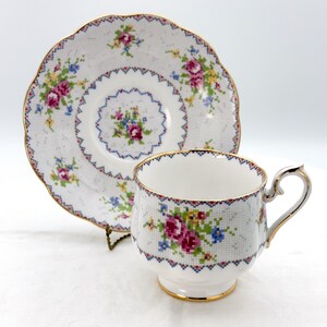 May include: A white teacup and saucer set with a floral cross-stitch pattern. The cup and saucer have gold trim and a scalloped edge. The floral design includes pink, blue, and yellow flowers.