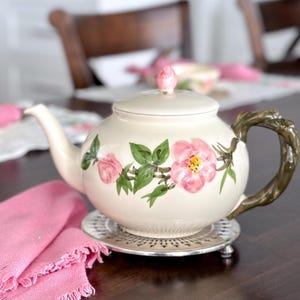 May include: Cream-colored ceramic teapot with a floral design featuring pink roses and green leaves. The teapot has a brown handle and sits on a silver trivet. A pink napkin is next to the teapot. Tea set.