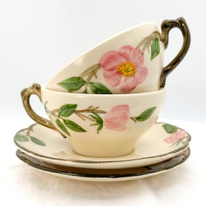 May include: Two stacked teacups and saucers with a floral design. The cups and saucers are cream-colored with pink flowers, green leaves, and brown stems. The handles are brown. The saucers are stacked on top of each other.