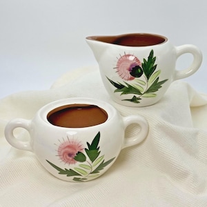 May include: A white ceramic creamer and sugar bowl set. Each piece features a hand-painted thistle design in pink, green, and purple. The creamer has a brown interior and a handle. The sugar bowl has a matching brown interior and two handles.