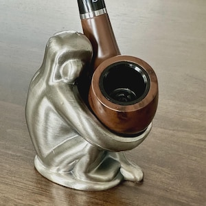 May include: A silver-colored sculpture of a person embracing a brown wooden pipe. The pipe has a black interior and a black mouthpiece. The sculpture is on a wooden surface.