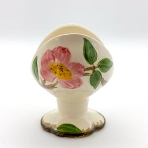 May include: A cream-colored ceramic napkin holder with a pedestal base and a scalloped edge. The front features a hand-painted design of a large pink flower with a yellow center and green leaves. The piece has a smooth, glossy finish throughout.