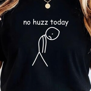 Op de afbeelding: Zwart T-shirt met de tekst "no huzz today" en een witte tekening van een stokfiguur. Het shirt heeft korte mouwen en wordt gedragen met een blauwe spijkerbroek en een bruine riem. Het Axis Shirt-logo is zichtbaar.
