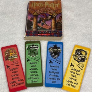 May include: A copy of "Harry Potter and the Sorcerer's Stone" with four house-themed bookmarks. The bookmarks are red, green, blue, and yellow, each with a house crest and values listed. The book cover features a wizard flying.