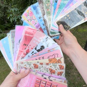 May include: A collection of rectangular bookmarks fanned out in a display. The bookmarks feature various designs including floral patterns, highland cows, bookshelves, and text such as 'I dust my shelves but my books are still filthy'.