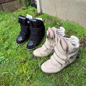 May include: Two pairs of wedge sneakers on green grass. One pair is black with three hook-and-loop straps, and the other is beige with three hook-and-loop straps. Both pairs have a high-top design.