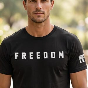 May include: Black crew neck t-shirt with the word "FREEDOM" printed in white across the chest. A small American flag is on the left sleeve. The shirt is made of a soft material.