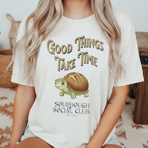 Op de afbeelding: Crèmekleurig T-shirt met de tekst "Good Things Take Time" in gouden letters. Daaronder een cartoon schildpad met een broodkorst als schild. Het shirt heeft ook de tekst "Sourdough Social Club" en "Slow Living".