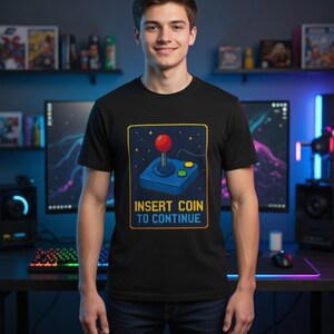 Könnte beinhalten: Schwarzes T-Shirt mit einem pixeligen Arcade-Joystick-Design, einschließlich einer roten Kugel und dem Text "INSERT COIN TO CONTINUE" in Gelb. Das Design ist in einem blauen und goldenen Rahmen gehalten. Die Person lächelt.