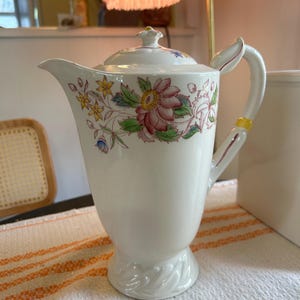 May include: White ceramic teapot with a lid, featuring a floral design in pink, yellow, and blue. The teapot has a curved handle and a spout for pouring. The base has a decorative swirl design. A classic piece for serving tea.