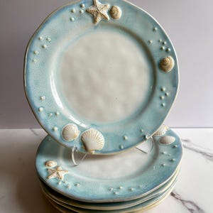 May include: A stack of four round ceramic plates featuring a white center and a light blue rim. The rim is decorated with raised, three-dimensional white starfish and seashell accents, along with small, scattered white dots resembling bubbles.