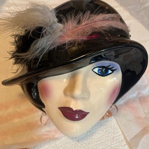 May include: A ceramic face sculpture with a black hat adorned with pink and gray feathers. The face has blue eyes, red lips, and blush on the cheeks. The sculpture has a glossy finish and is likely a decorative item.