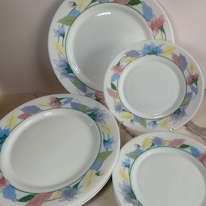 May include: A set of white ceramic plates featuring a decorative border of pastel-colored floral and leaf illustrations in shades of blue, pink, and yellow. The collection includes larger dinner plates and smaller salad plates with a smooth finish.