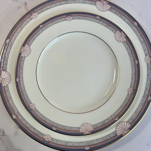 May include: Two white ceramic plates with a seashell design. The plates have a gold rim and a border with gray, blue, and pink seashell motifs. The plates are stacked on top of each other.
