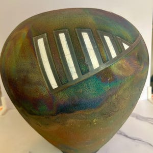 May include: A handcrafted, iridescent ceramic vase with a unique shape. The vase features a geometric design of white rectangles and a triangle on a dark background with shades of green, brown, and blue. The vase has a textured surface and a dark base.