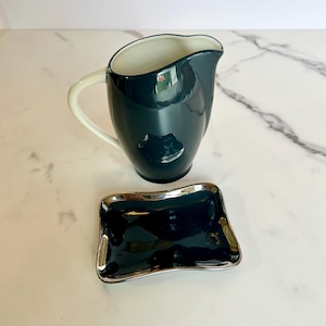 May include: A dark teal ceramic pitcher with a cream-colored handle and interior, next to a matching rectangular dish with a silver rim. The pitcher and dish have a glossy finish, reflecting light. The set is likely for serving.