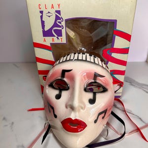 May include: A ceramic mask with a musical theme, featuring a white face with red lips, blush, and musical notes. The mask has a piano key design on the forehead and is accompanied by a box with the text "CLAY ART". Ribbons in various colors are attached.