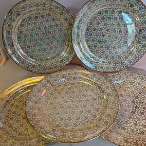 May include: A set of five decorative plates with a floral pattern. The plates have a gold rim and a pearlescent finish. The floral pattern is a mix of white, green, and gold. The plates are arranged in a circular pattern.
