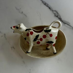 May include: A ceramic creamer shaped like a cow with a cream-colored base, speckled texture, and a red cherry pattern with green leaves. The cow-shaped pitcher features a curved handle, a spout at the mouth, and stands inside a matching oval bowl.