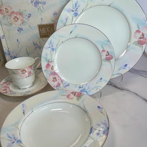 May include: A Mikasa floral dinnerware set featuring a teacup and saucer, plates, and a bowl. The white porcelain pieces are decorated with pink and blue floral patterns and gold rims. The set is displayed with its original box.