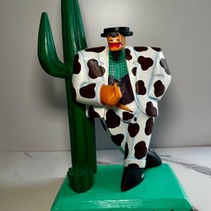 May include: A hand-painted wooden figurine of a figure wearing a white suit with black cow-print spots, a black hat, and a green checkered shirt, leaning against a tall green cactus on a rectangular green base with a decorative triangular border.