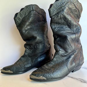 May include: A pair of black cowboy boots with a textured, crocodile-like pattern. The boots have a tall shaft, pointed toe, and decorative metal accents on the toe. The leather appears worn, giving them a vintage look.