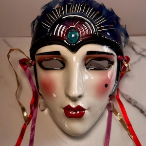 May include: A ceramic face mask featuring a glossy white finish with painted red lips, pink cheeks, and a black beauty mark. The mask is adorned with a dark blue headpiece, blue feathers, a central teal gem, and dangling ribbons in red, gold, and purple.