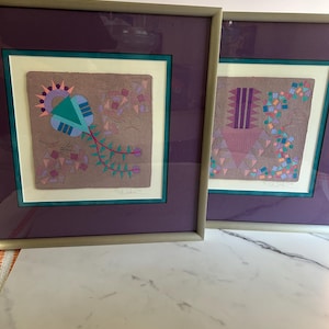 May include: Two framed abstract art prints. Each print features a colorful geometric design on a textured background, framed in a silver-tone frame with a purple mat. The artwork includes shapes like triangles, rectangles, and floral elements.