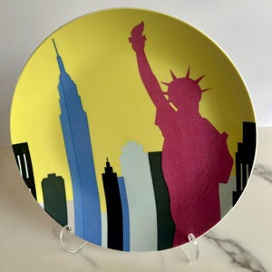 May include: A decorative plate featuring a yellow background with a New York City skyline design. The plate includes a pink silhouette of the Statue of Liberty and a blue silhouette of the Empire State Building.