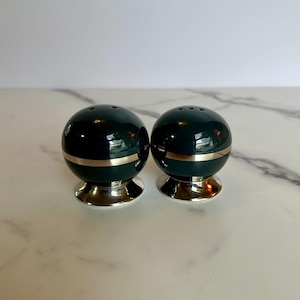 May include: Two dark green salt and pepper shakers with a gold band and silver base. Each shaker has three holes on top for dispensing. The shakers are spherical in shape.