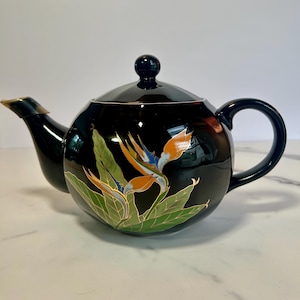 May include: Black ceramic teapot with a lid and handle. The teapot features a colorful floral design of a bird of paradise flower with orange, blue, and green accents. The spout has a gold-colored trim.