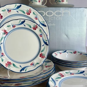 May include: A complete Boch by Mikasa dinnerware set featuring white plates, bowls, cups, and saucers. Each piece is decorated with a blue rim and floral pattern in red and green. The set includes multiple plates, bowls, cups, and saucers.